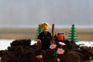 Picture of Lego figure in suit with briefcase holding shovel standing on soil. Lego flowers are blooming out of them.