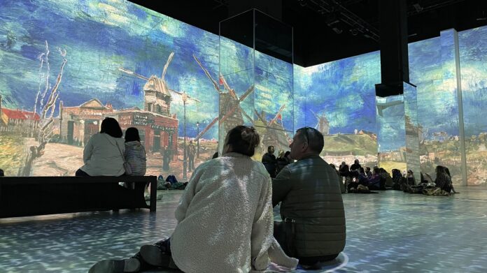 A couple sitting on the floor while staring at two painted windmills in the Immersive Van Gogh exhibition